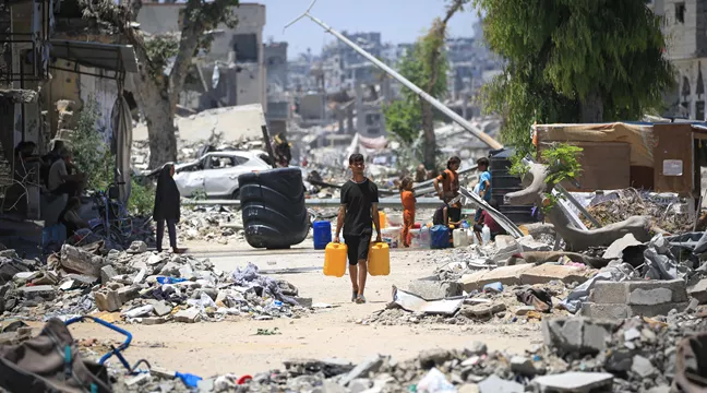 En palestinsk man bär två vattendunkar förbi förstörda byggnader i Khan Yunis, i södra Gaza, foto. 