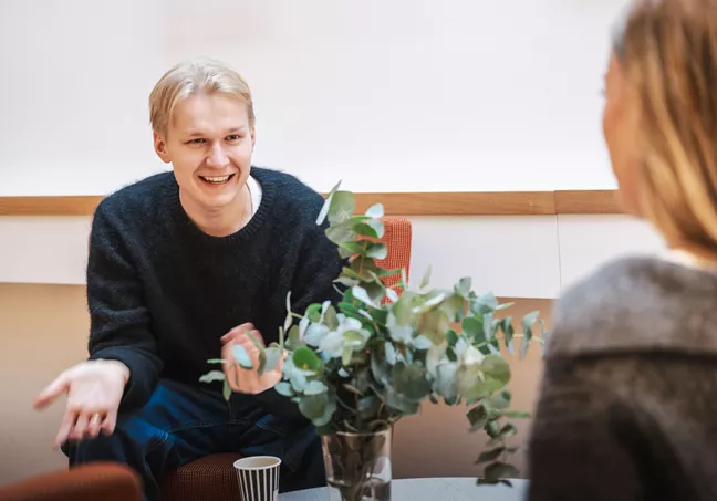 Emil sitter och pratar med en annan person som man bara ser ryggen på. Han ser glad ut och gestikulerar. 