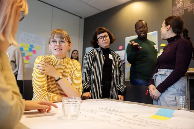 Fem personer står och sitter runt ett bord. De ser ut att vara involverade i djupa samtal. 